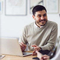 Caucasian businessman is sitting in an office and having a meeting with a female colleague. He might be working from home. It's morning or later in the day. He is smiling. A laptop is in front of him and the woman is holding a digital tablet.